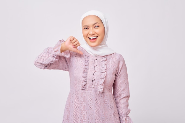 Portrait de souriante belle jeune femme musulmane asiatique portant le hijab et la robe violette fière d'elle-même tout en pointant le doigt sur lui-même isolé sur fond de studio blanc