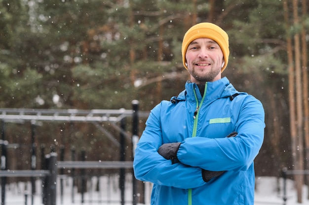 Portrait de sourire jeune athlète masculin en veste chaude debout avec les bras croisés sous la neige qui tombe à la zone d'entraînement