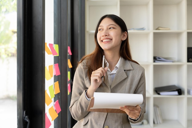 Portrait sourire jeune femme asiatique prenant des décisions en pensant à la fenêtre du bureau