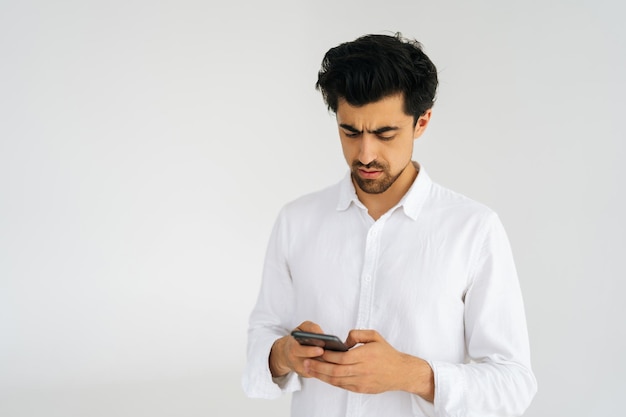 Portrait en studio de beau jeune homme concentré en chemise tenant un smartphone regardant sur l'écran de l'appareil debout sur fond isolé blanc