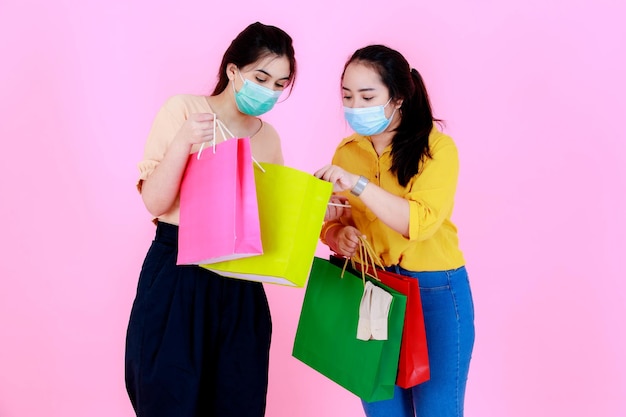 Portrait en studio de deux jeunes amies asiatiques heureuses d'acheteurs portant un masque facial et une tenue décontractée portant des sacs à provisions colorés ensemble pendant la quarantaine pandémique covid sur fond rose.