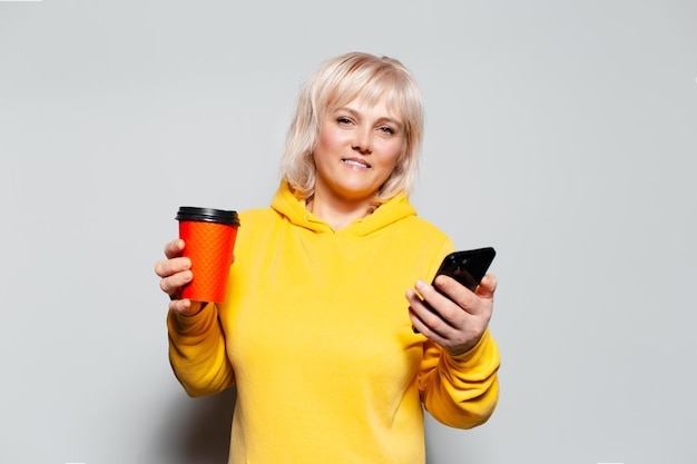 Portrait en studio d'une femme blonde à l'aide d'un smartphone tenant une tasse de papier pour le café à emporter de couleur rouge portant un pull jaune sur fond blanc