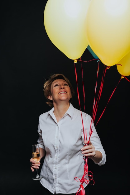Portrait en studio de femme tenant un verre de champagne et des ballons jaunes souriant loin isolé sur fond noir