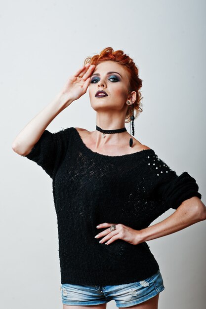 Portrait en studio de la fille aux cheveux rouge sur une blouse noire et un short en jean avec un maquillage foncé et brillant