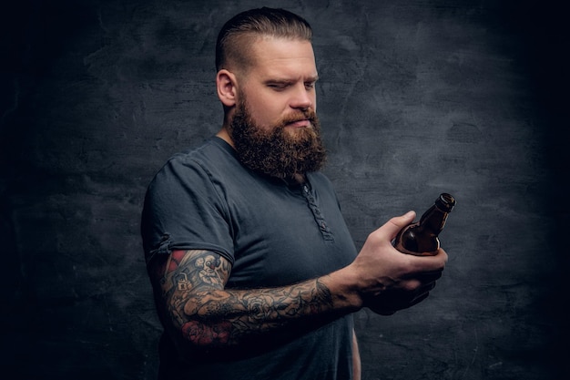 Portrait en studio d'un hipster barbu brutal avec tatouage sur le bras, regardant la bouteille de bière sur fond de vignette grise.