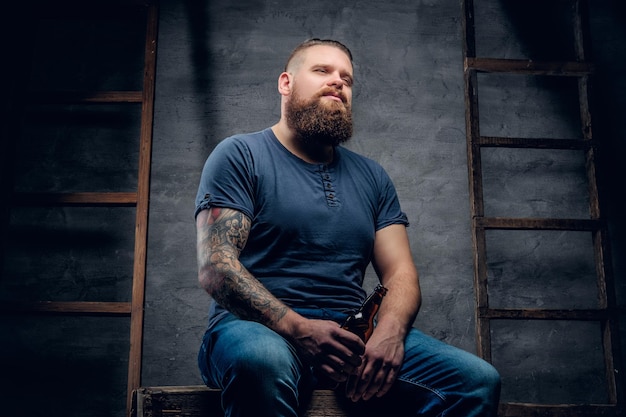 Portrait en studio d'un hipster tatoué barbu tenant la bouteille de bière sur fond gris avec deux échelles en bois vintage.