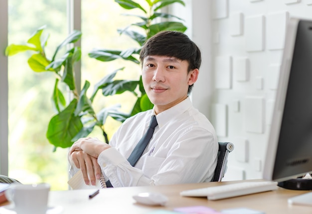 Portrait en studio d'un homme d'affaires professionnel asiatique prospère en chemise formelle avec cravate assis regardant la caméra au bureau avec écran d'ordinateur, clavier, souris et papeterie.