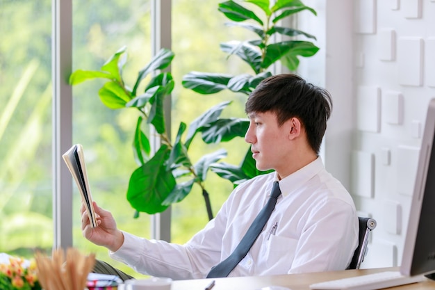 Portrait en studio d'un homme d'affaires professionnel asiatique prospère en chemise formelle avec cravate assis regarder la caméra au bureau avec écran d'ordinateur clavier souris et papeterie