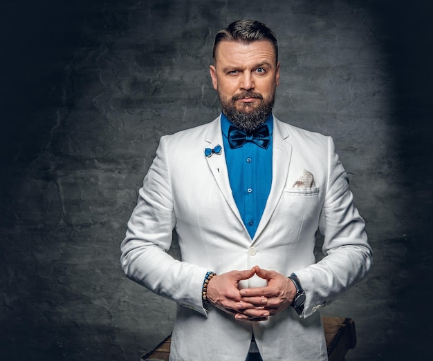 Portrait en studio d'un homme barbu vêtu d'une chemise bleue, d'une veste blanche et d'un nœud papillon sur fond gris.