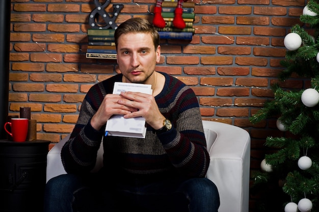 Portrait en studio d'un homme avec un livre assis sur une chaise contre un arbre de noël avec des décorations.