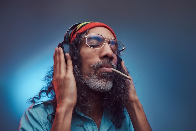 Le portrait en studio d'un homme rastafarien africain aime la musique dans les écouteurs et fumer de l'herbe. Isolé sur un fond bleu.