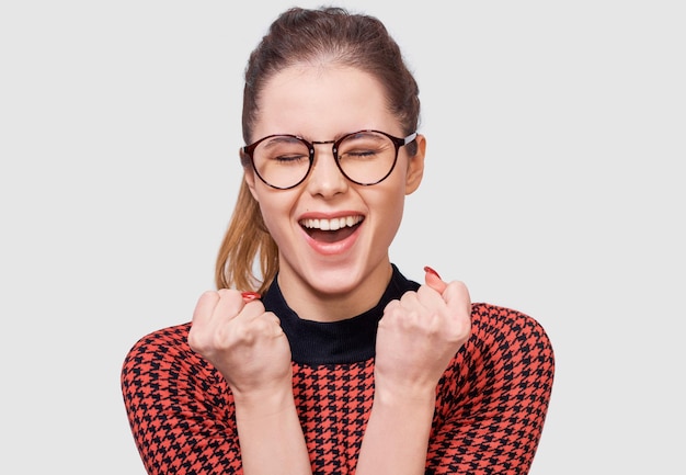 Portrait en studio horizontal d'une jeune femme aux yeux fermés, les poings serrés, se réjouit du succès portant des lunettes rondes transparentes posant sur fond blanc