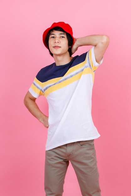 Portrait en studio d'un jeune asiatique beau adolescent urbain modèle masculin de mode en tenue de rue t-shirt sport pantalon beige et chapeau de seau rouge debout posant regarder la caméra sur fond rose