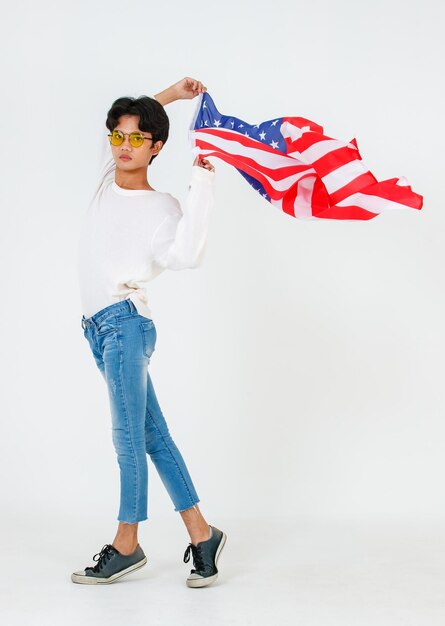 Portrait en studio d'un jeune asiatique LGBT gay bisexuel homosexuel masculin modèle à la mode en tenue décontractée et lunettes de soleil de mode debout tenant en agitant le drapeau national des États-Unis sur fond blanc