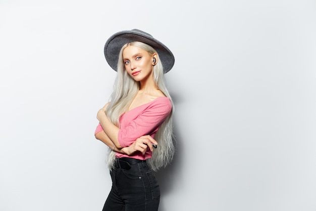 Portrait en studio de jeune belle fille blonde avec des écouteurs sans fil à l'oreille portant un chapeau gris et une chemise rose sur fond blanc Espace de copie