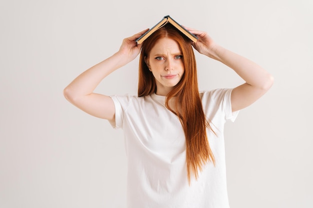 Portrait en studio d'une jeune étudiante en colère couvrant la tête avec un livre à l'écart debout sur blanc