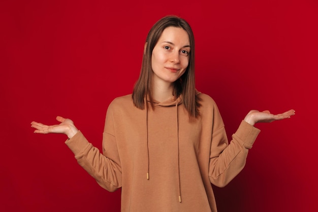 Portrait en studio d'une jeune femme haussant les épaules indécise sur fond rouge