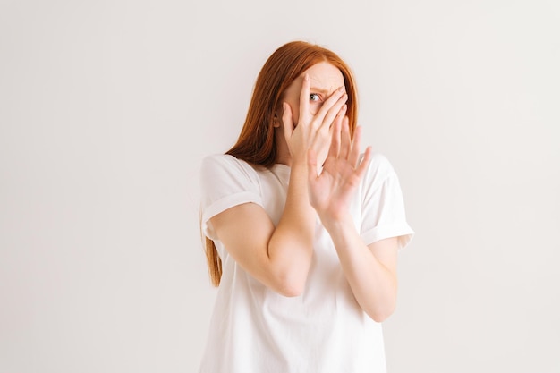 Portrait en studio d'une jeune femme rousse effrayée à l'air timide ou terrifiée couvrant le visage caché avec les mains lorgnant à travers les doigts choqué par un film d'horreur ou effrayé sur fond blanc isolé