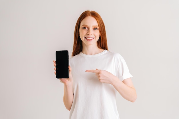 Portrait en studio d'une jeune femme rousse joyeuse avec un large sourire tenant un téléphone portable à écran blanc et pointant l'index, regardant la caméra, debout sur un fond blanc isolé.