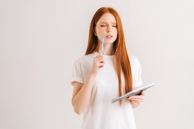 Portrait en studio d'une jeune femme rousse réfléchie, les yeux fermés, toucher le visage avec un stylo tenant un journal debout sur un fond isolé blanc Réfléchir à une étudiante prenant des notes dans un cahier papier