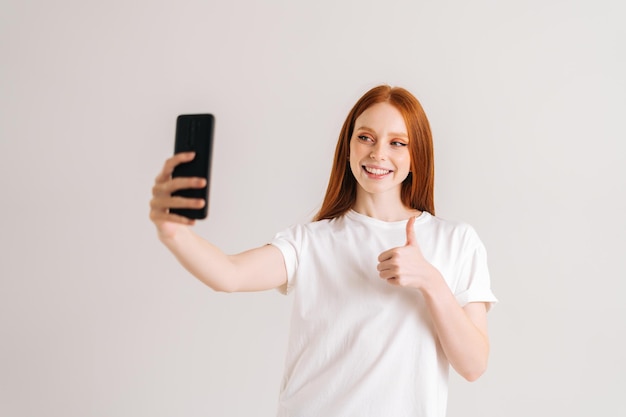 Portrait en studio d'une jeune femme souriante prenant un selfie et montrant les pouces vers le haut à l'aide d'un smartphone debout