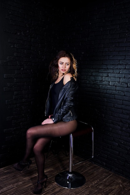 Portrait en studio de jeune fille brune sexy en veste de cuir noir contre le mur de briques