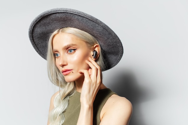 Portrait en studio de jeune fille séduisante blonde avec des écouteurs dans les oreilles sur fond blanc portant un chapeau gris Toucher l'écouteur sans fil