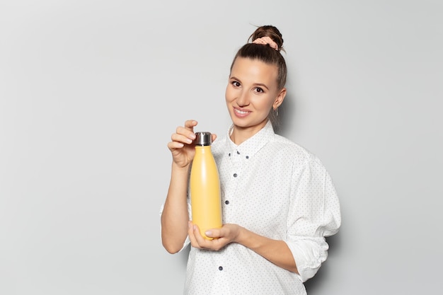 Portrait en studio d'une jeune fille souriante ouvre la bouteille thermo de couleur jaune sur fond blanc