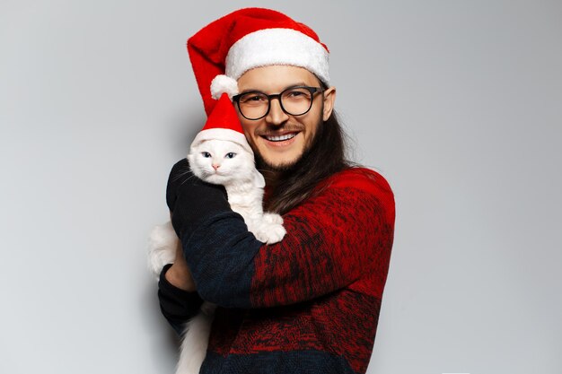 Portrait en studio d'un jeune homme de bonheur porte un pull de Noël rouge étreint un chat blanc portant un bonnet de Noel sur fond blanc