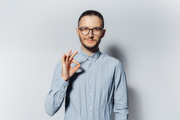 Portrait en studio d'un jeune homme confiant montrant un geste ok sur fond blanc portant une chemise bleue et des lunettes