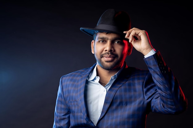 Portrait en studio d'un jeune homme indien mal rasé et confiant, homme d'affaires portant une chemise, un costume à carreaux, regardant la caméra, touchant un chapeau, souriant, debout isolé sur fond sombre.