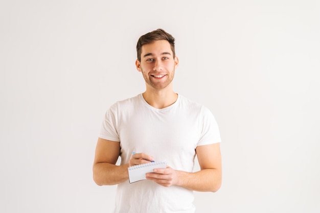 Portrait en studio d'un jeune homme joyeux écrivant dans un cahier avec un stylo debout sur un fond isolé blanc