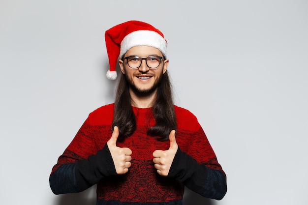 Portrait en studio d'un jeune homme souriant aux cheveux longs et aux lunettes montrant le geste du pouce levé porte un pull de Noël rouge et un bonnet de Noel sur fond blanc