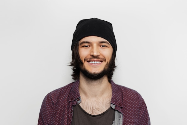 Portrait en studio de jeune homme souriant avec un chapeau noir sur une surface blanche.