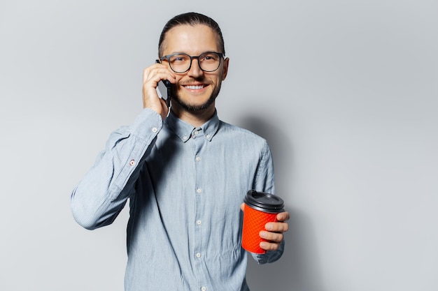 Portrait en studio d'un jeune homme souriant parlant sur un smartphone tenant une tasse de papier rouge pour le café à emporter dans une autre main sur fond blanc portant une chemise bleue et des lunettes