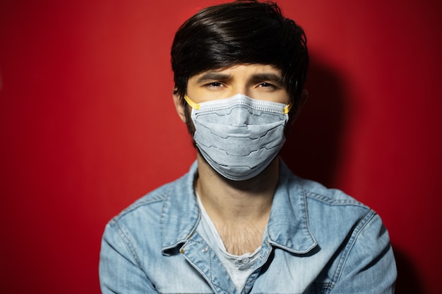 Portrait en studio de jeune homme en veste en jean portant un masque médical contre le coronavirus sur une surface rouge.