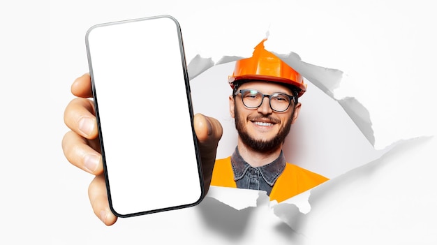 Portrait en studio d'un jeune ouvrier du bâtiment souriant montrant un gros smartphone avec un blanc à l'écran à travers un trou de papier déchiré Fond blanc Portant des lunettes et un équipement de sécurité