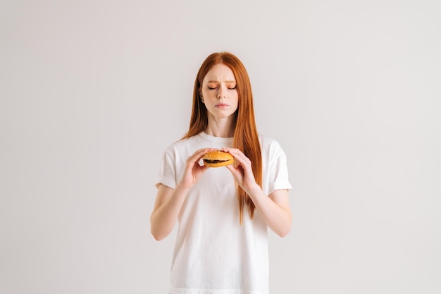 Portrait en studio de jolie jeune femme mécontente avec dégoût tenant un hamburger regardant vers le bas debout sur fond isolé blanc Triste jolie femme rousse mangeant un hamburger