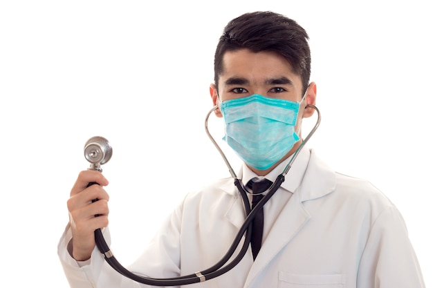 Portrait en studio d'un médecin de sexe masculin en uniforme et masque avec stéthoscope dans ses mains posant isolé sur mur blanc