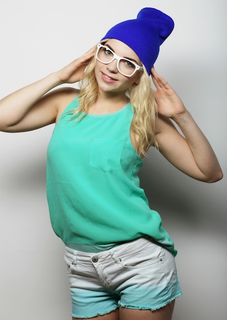 Portrait en studio de mode d'une jolie jeune femme blonde hipster avec des lunettes, portant un t-shirt urbain élégant et un chapeau, sur fond blanc
