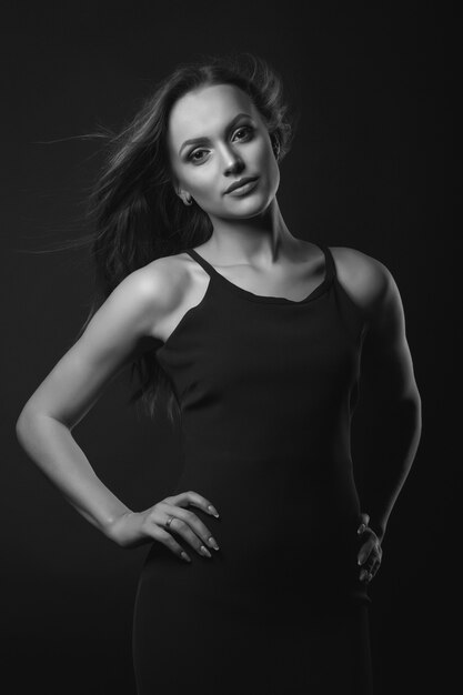 Portrait en studio de mode d'une superbe femme brune avec un maquillage et une coiffure parfaits porte une robe noire classique posant dans un studio sombre. Prise de vue en noir et blanc