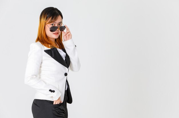 Portrait en studio d'un modèle de femme d'affaires à la mode et à la mode urbaine asiatique en costume de blazer décontracté regarde la caméra en ajustant les lunettes de soleil aviateur à lentille noire sur fond blanc.