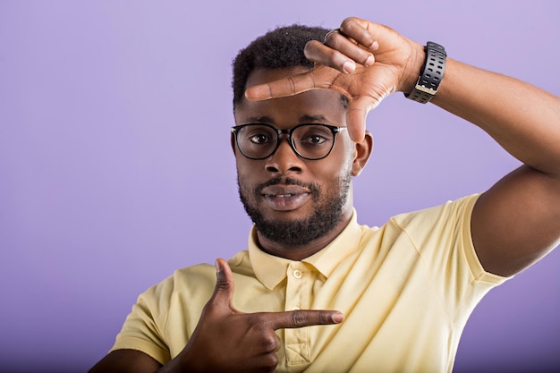 Portrait en studio d'un modèle masculin afro-américain joyeux et insouciant faisant un geste de cadre avec les doigts devant son visage, regardant la caméra, souriant largement sur fond lilas.