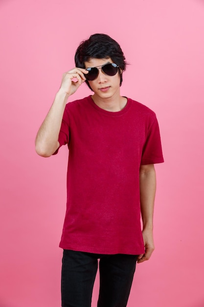 Portrait en studio d'un modèle masculin de mode asiatique jeune adolescent urbain en tenue de rue décontractée t-shirt et jeans noirs avec des lunettes de soleil aviateur debout posant regarder la caméra sur fond rose