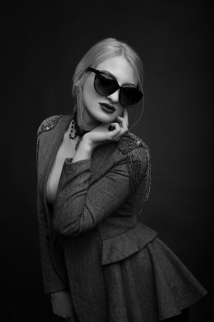 Portrait en studio noir et blanc d'une fabuleuse femme blonde porte des lunettes de soleil et une veste sur un corps nu