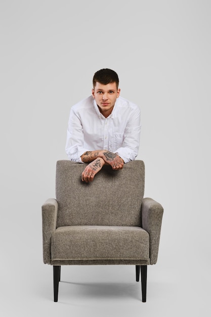 Portrait en studio de pleine longueur d'un jeune homme aux pieds nus en chemise blanche et pantalon ivoire se penchant sur un fauteuil