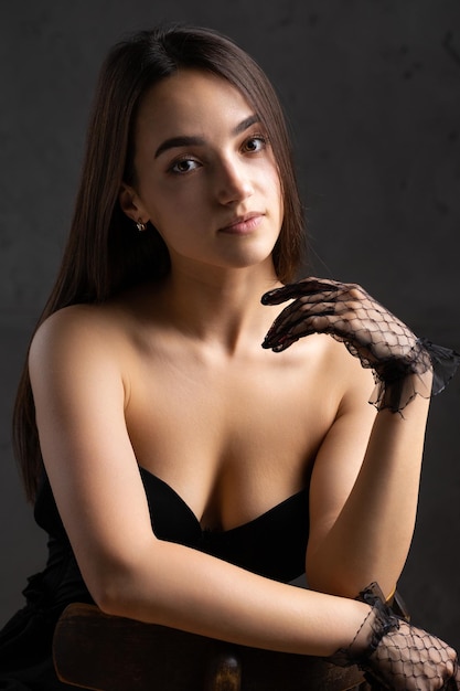 Portrait de studio sombre classique d'une jeune femme brune vêtue de noir qui est assise sur une chaise