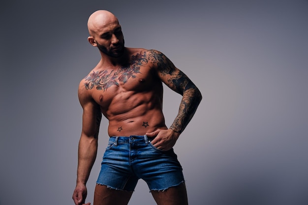 Portrait en studio de tête rasée torse nu, homme musclé avec des tatouages sur son torse sur fond de vignette grise.