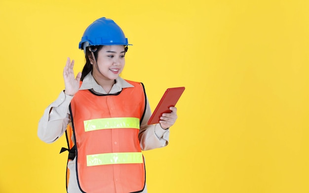 Portrait studio tourné femme asiatique contremaître d'ingénierie professionnel en casque dur et gilet de sécurité réfléchissant souriant regarder la caméra vérifiant le stock d'inventaire de la tablette sur fond jaune.