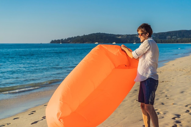 Portrait de style de vie d'été de l'homme gonfle un canapé orange gonflable sur la plage de l'île tropicale Se détendre et profiter de la vie sur un lit d'air